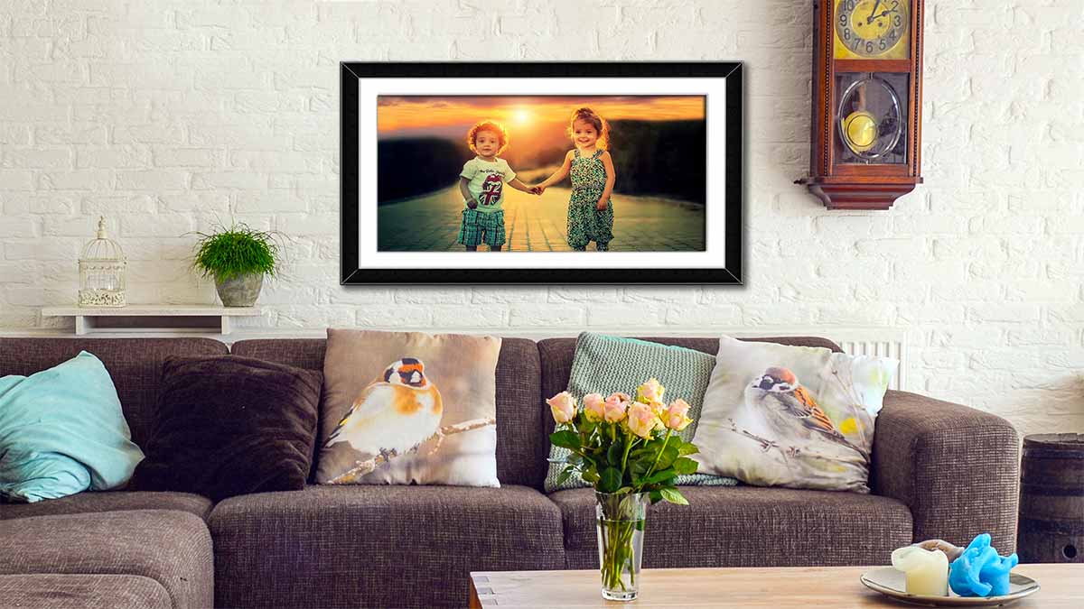 Picture of two children holding hands in the sunset, framed poster in a simple black frame hanging over a sofa with decorative cushions