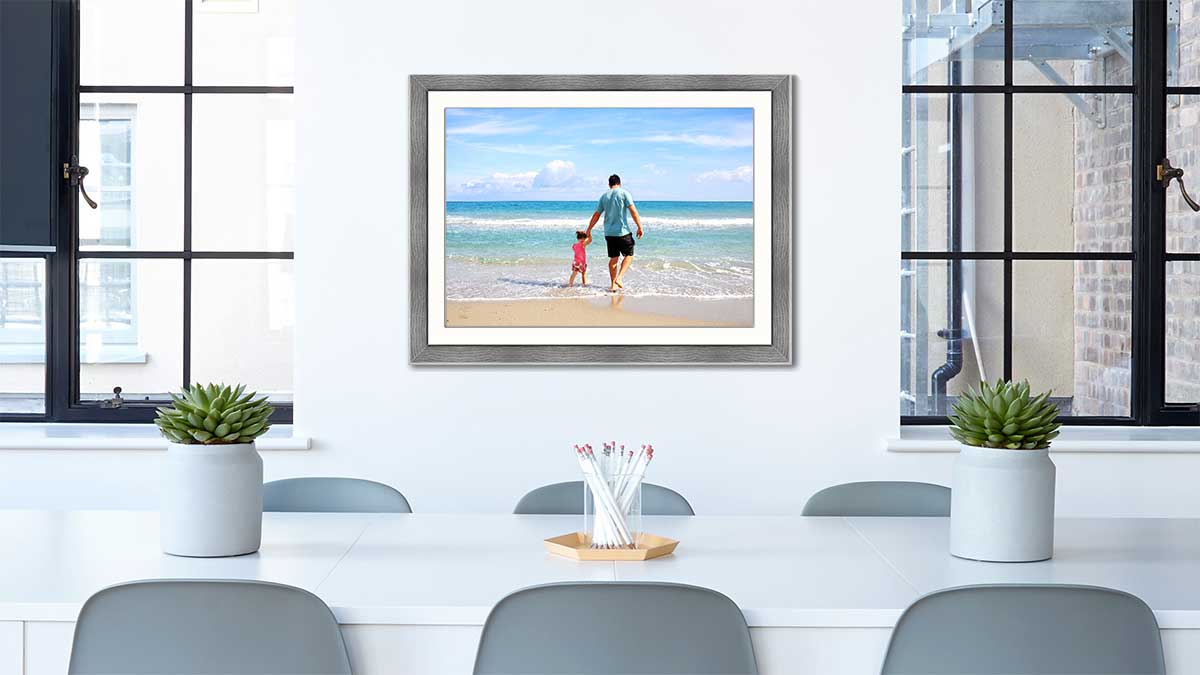Family picture of a father with child playing in the water at the seaside. Framed in a grey wood frame and hung in a dinning room behind a white table with grey chairs