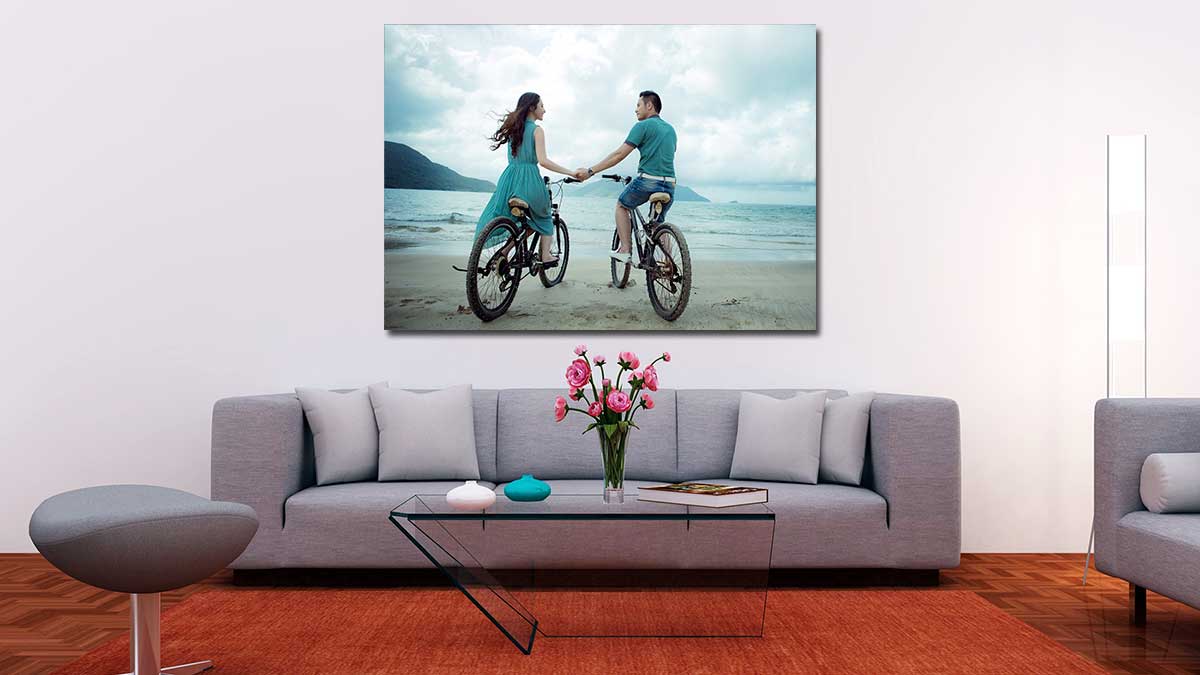 A1 poster featuring a romantic image of a couple riding bicycles on the beach and holding hands. Poster print is hung in a sitting room above a large sofa.