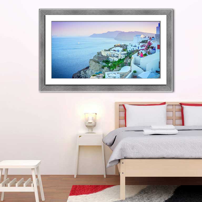 Sunset holiday snap of Santorini in Greece, framed in a grey open grain frame and hung in a minimalist styled bedroom with grey sheets on the bed and a grey rug.