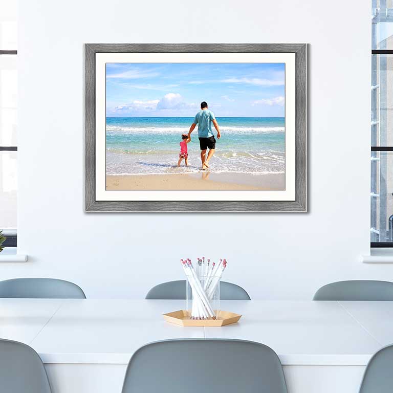 Family picture of a father with child playing in the water at the seaside. Framed in a grey wood frame and hung in a dinning room behind a white table with grey chairs